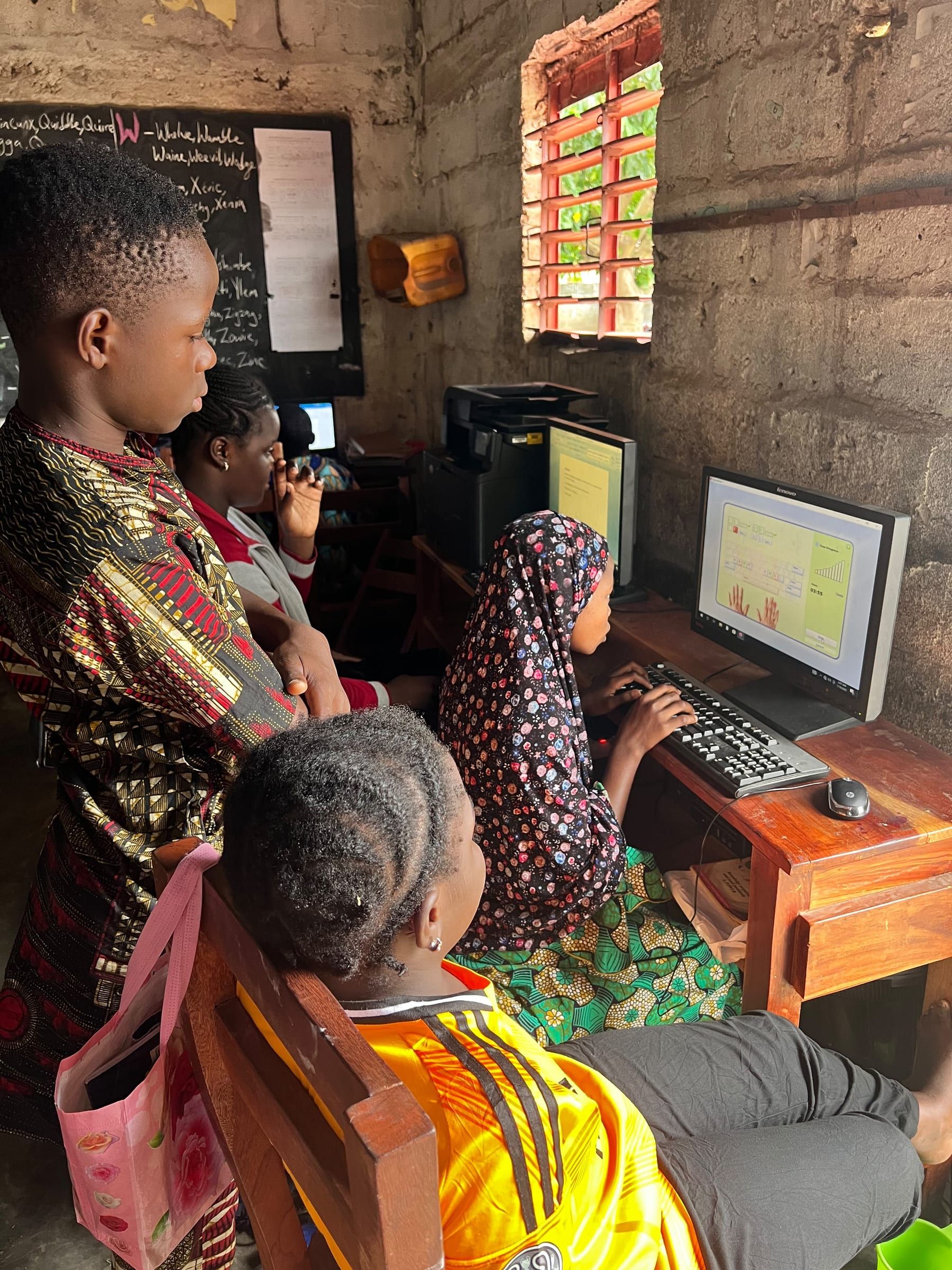 Des jeunes réunis autour d’un ordinateur dans la salle informatique de l’école, l’un d’eux au clavier ; derrière eux, un second écran, une imprimante et un tableau noir.