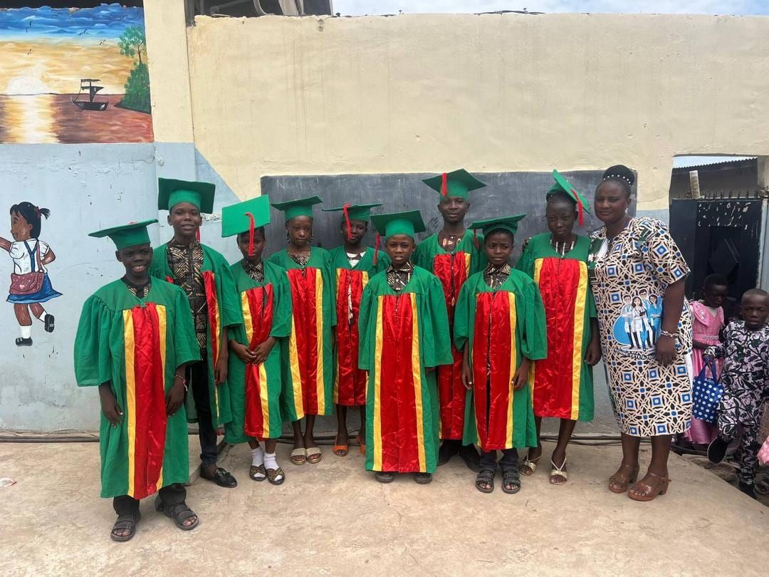 Des élèves en toge verte, avec écharpes rouge et jaune et chapeaux de diplômé, entourés de deux adultes devant un mur peint.