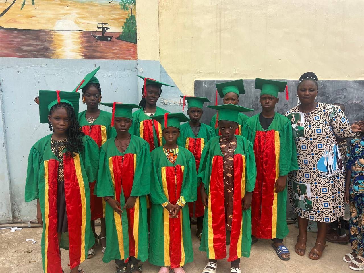 Des élèves en toge verte, avec écharpes rouge et jaune et chapeaux de diplômé, réunis avec une adulte devant un mur peint.