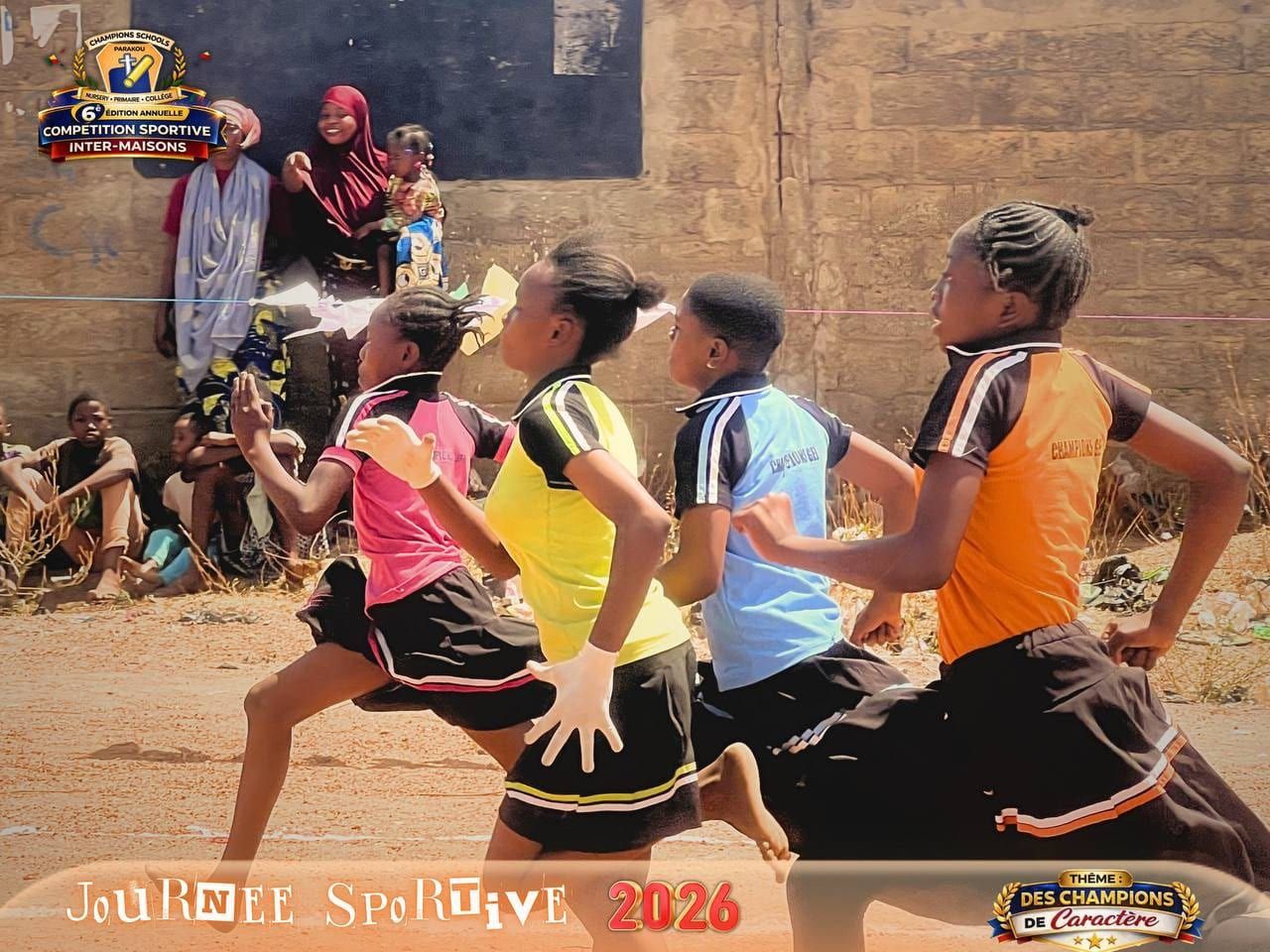 Quatre filles en tenue de maison rose, jaune, bleu clair et orange courent une course sur un terrain sablonneux, des spectateurs assis contre un mur derrière elles. La photographie porte le cadre « Journée Sportive 2026 » de l’école.